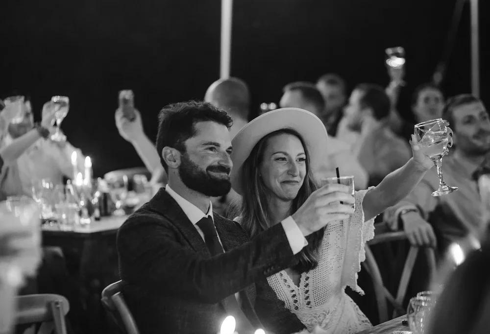 Couple raising glasses during wedding reception toast with guests reacting in the background