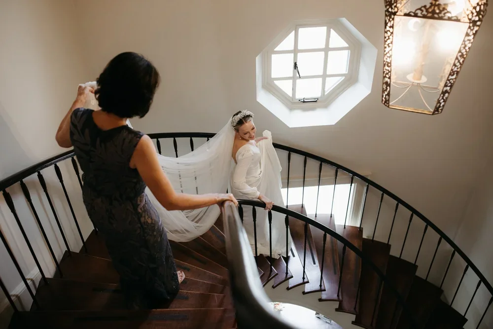 Bride descending staircase with mother adjusting veil during wedding morning getting ready coverage