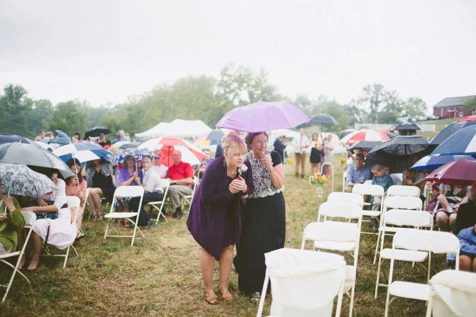 Candid wedding photo of guests under umbrellas during rainy outdoor ceremony, real emotional wedding moment