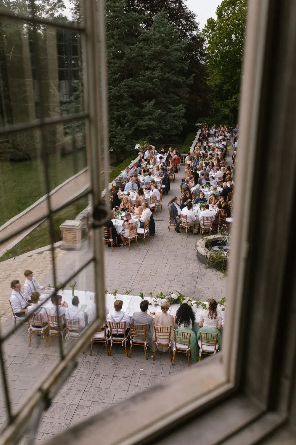 Birds-eye view of outdoor wedding reception seen through window frame with long tables and guests dining.