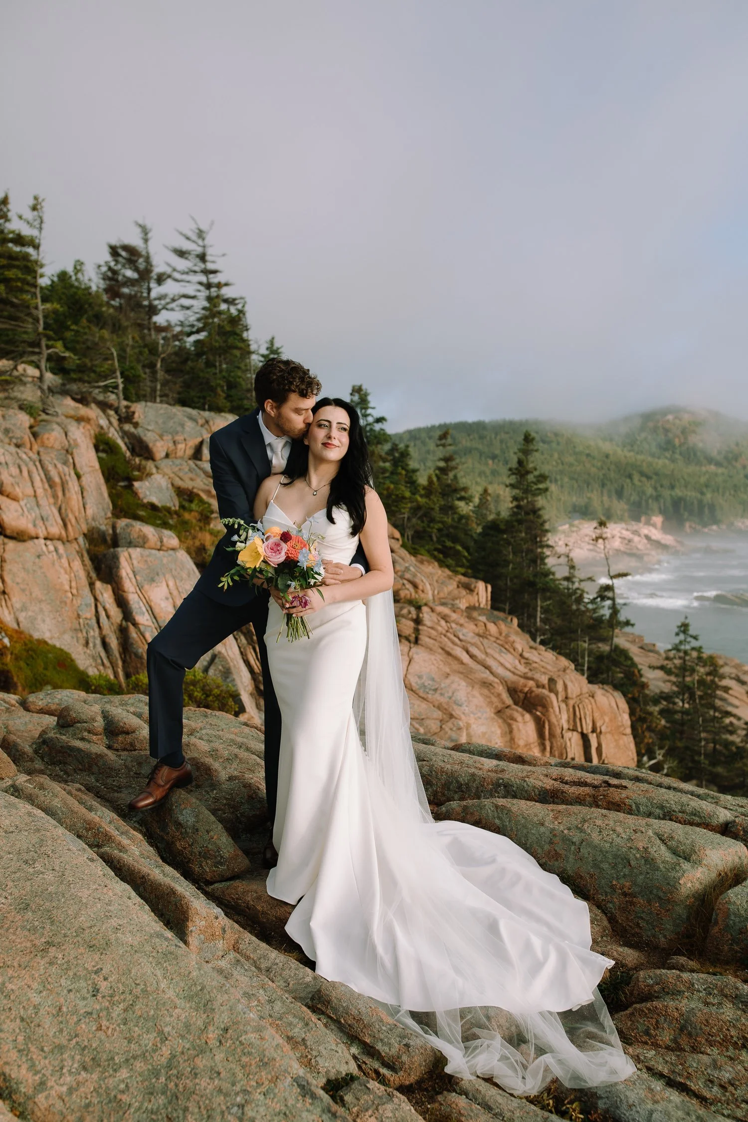 Couple standing together on coastal cliffs during an Acadia National Park elopement surrounded by ocean and evergreens.