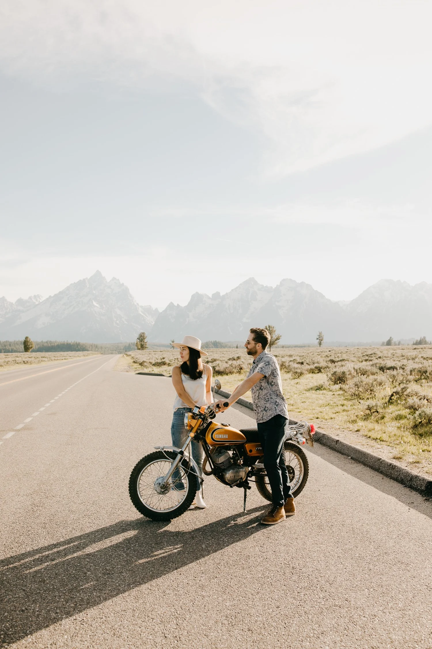 Engaged couple leaning against a motorcycle with the Teton Range in the background