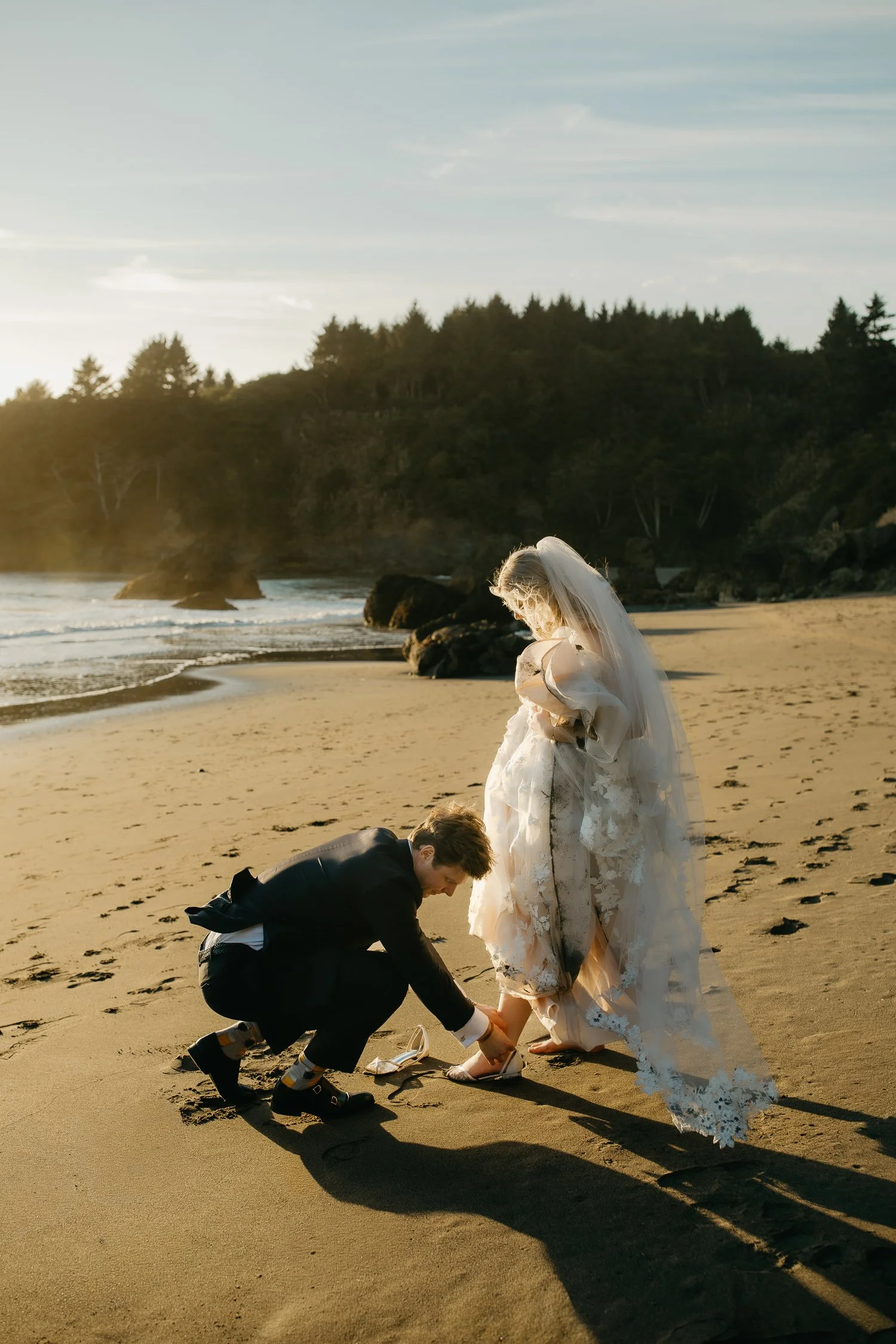 groom helping bride with shoe at sunset on the Northern California coast