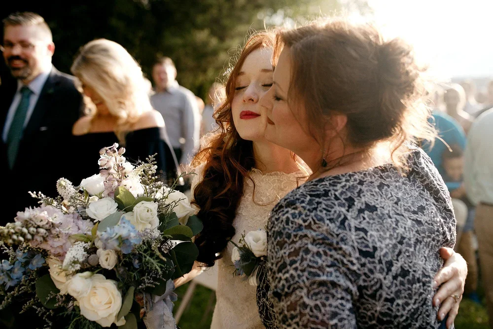 Bride embracing her mother in warm golden light after the outdoor wedding ceremony while holding her bouquet of white and pastel flowers.