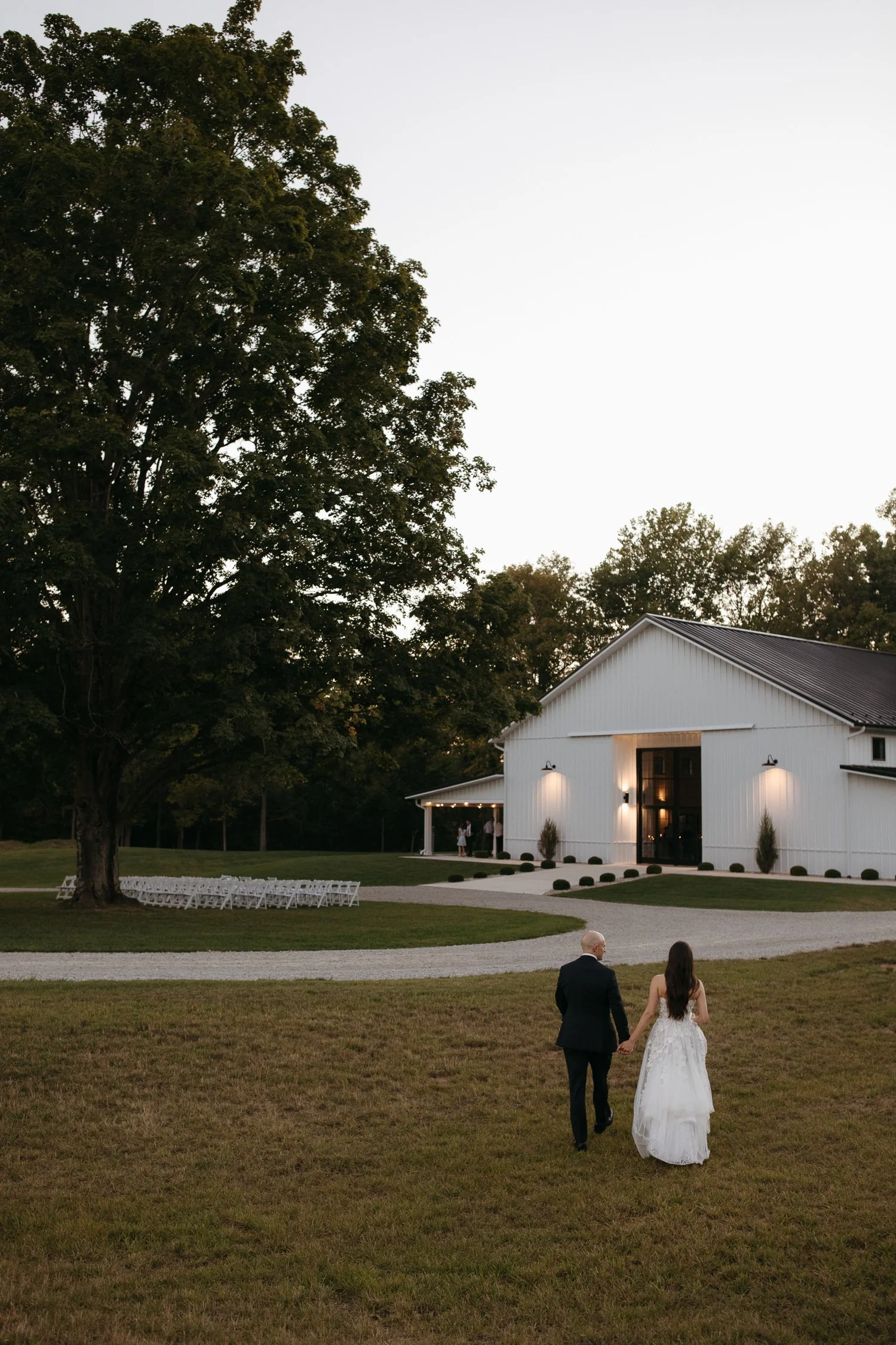 Bride and groom walking together across the lawn toward a white barn wedding venue at sunset