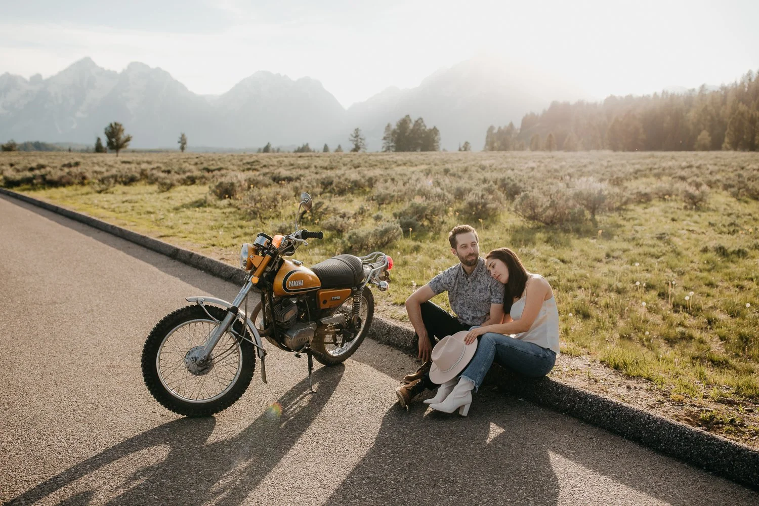 Wide view of couple sitting beside a motorcycle with the Teton Range in the background