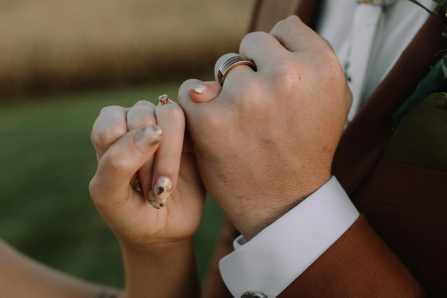Close-up of wedding rings as a bride holds the groom’s hand, featuring a rust-colored suit during an Ivory Meadows wedding in Dayton, Ohio.
