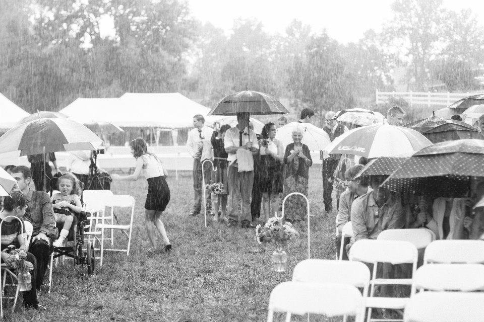 Black and white photo of rainy outdoor wedding ceremony with guests holding umbrellas, meaningful candid moment
