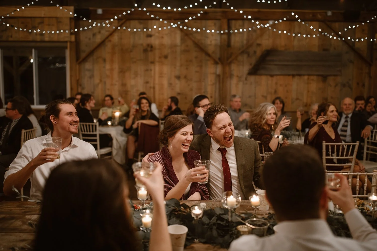 Wedding guests laughing and raising glasses during a reception toast in a warmly lit barn venue