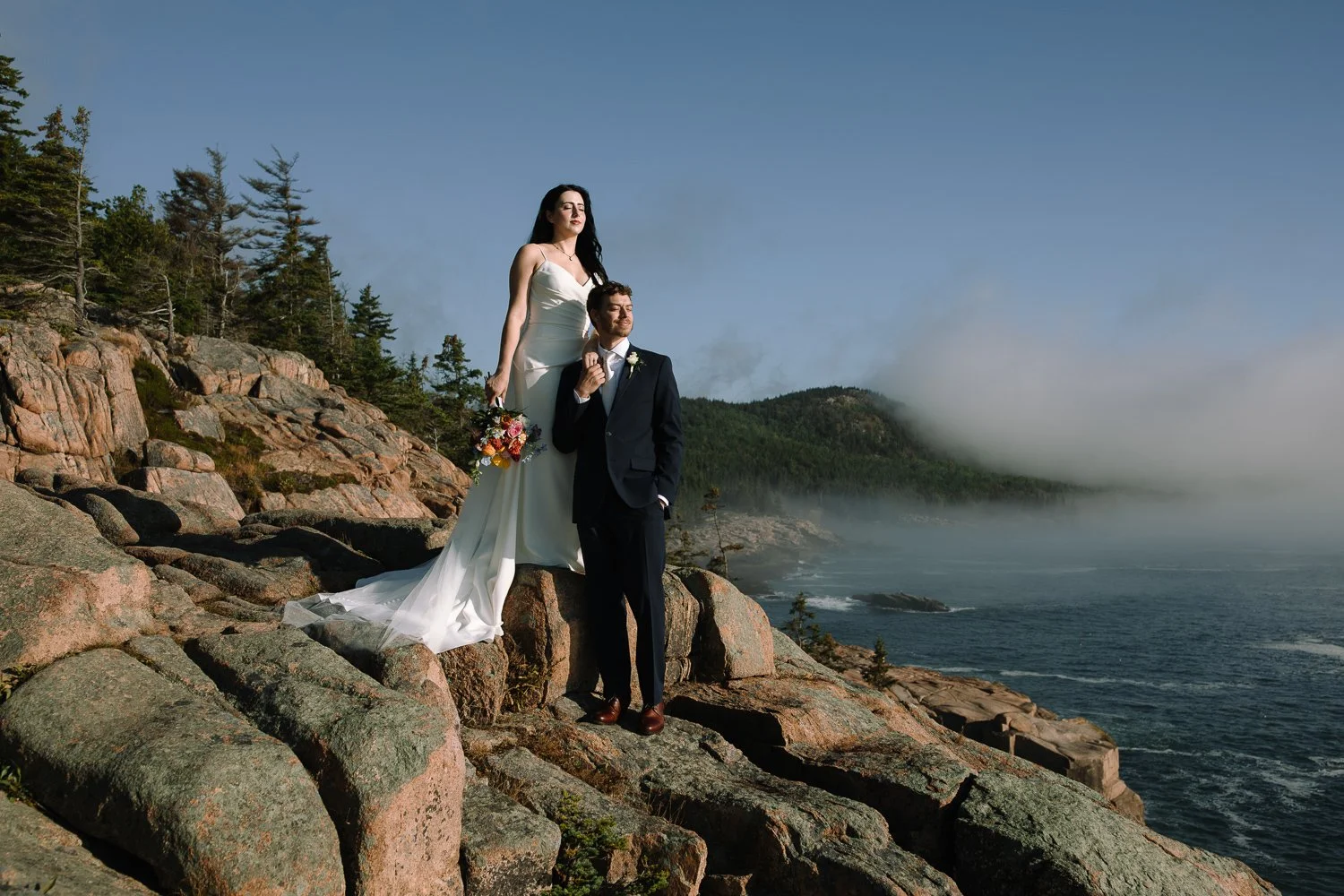 Couple standing on rocky cliffs as fog lifts during an early morning Acadia National Park elopement