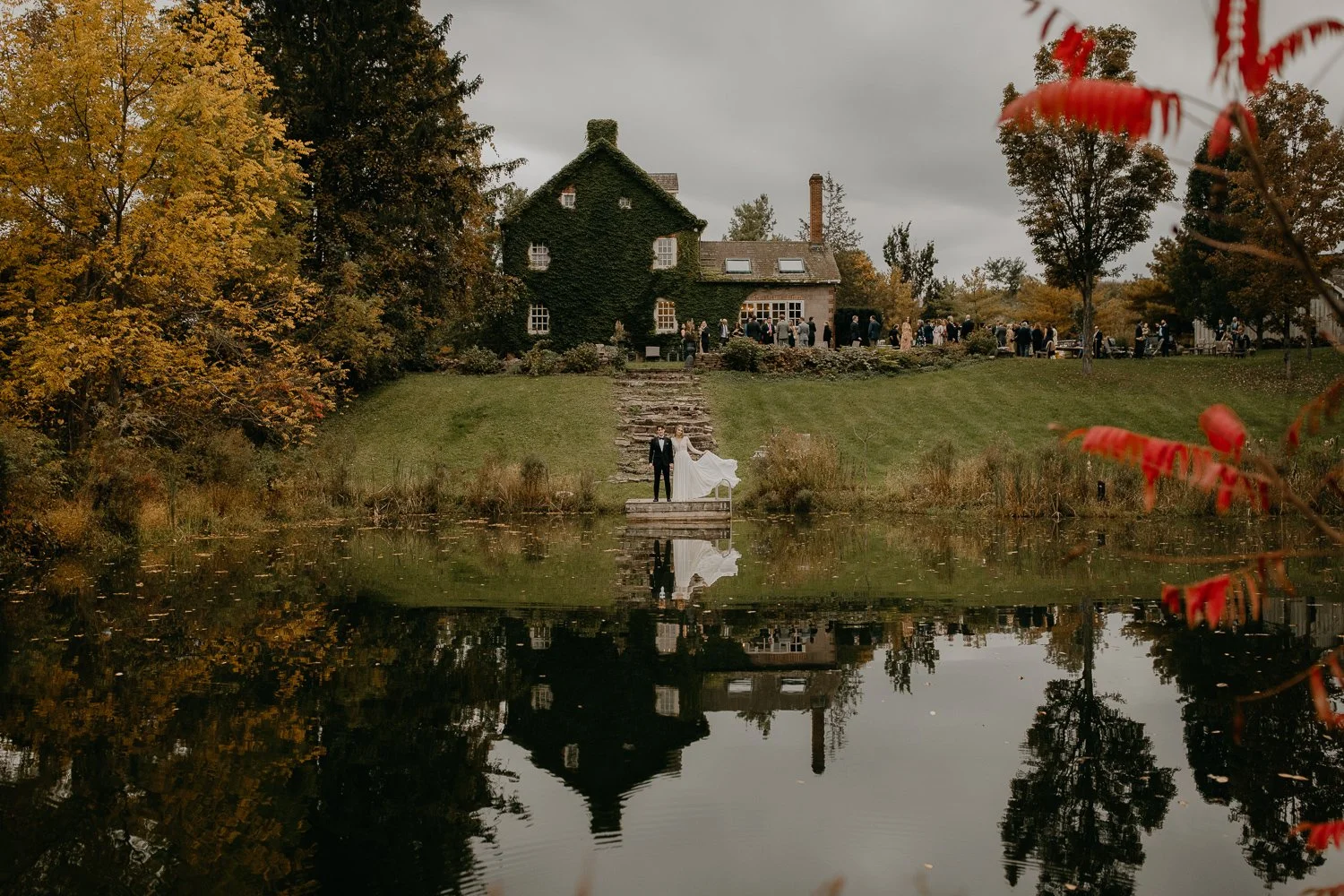 Wide autumn wedding portrait at estate with couple reflected in pond and guests gathered near ivy-covered house