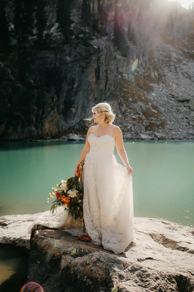Bride standing on a rocky lakeshore with turquoise water and mountain cliffs in Grand Teton National Park.