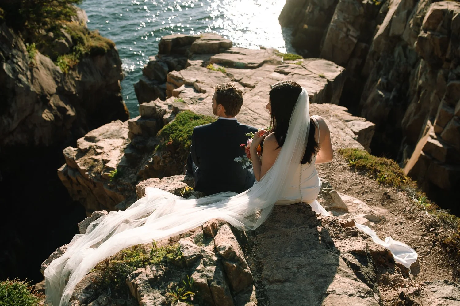 Bride and groom sitting together on rocky cliffs overlooking the ocean during a quiet Acadia elopement at sunset