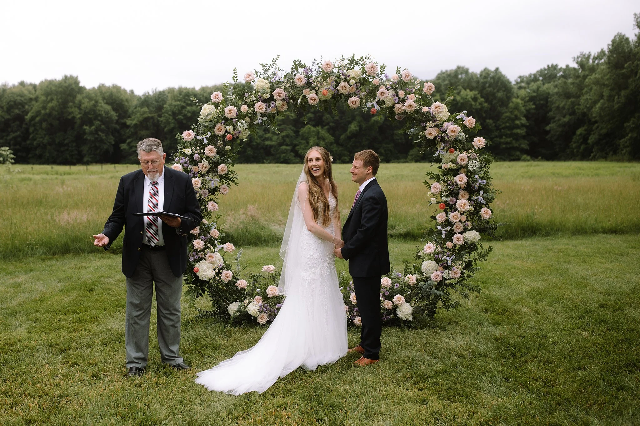 Bride and groom holding hands during an outdoor wedding ceremony beneath a floral arch in an open field.