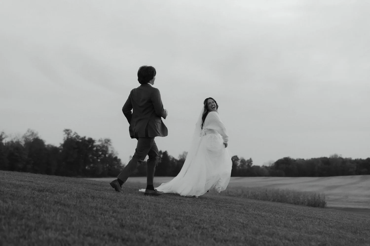 Candid black and white photo of a bride and groom joyfully running up a grassy hillside after their wedding ceremony.