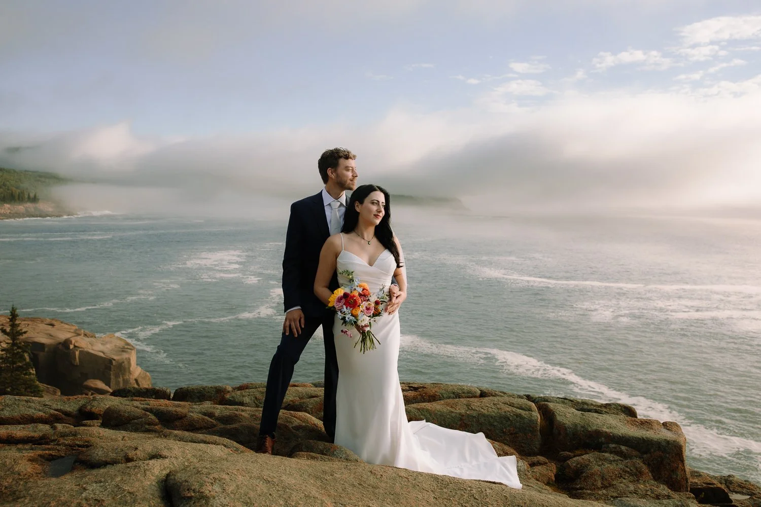 Bride and groom standing on the rocky coastline during an intimate Acadia National Park wedding with morning fog lifting over the ocean
