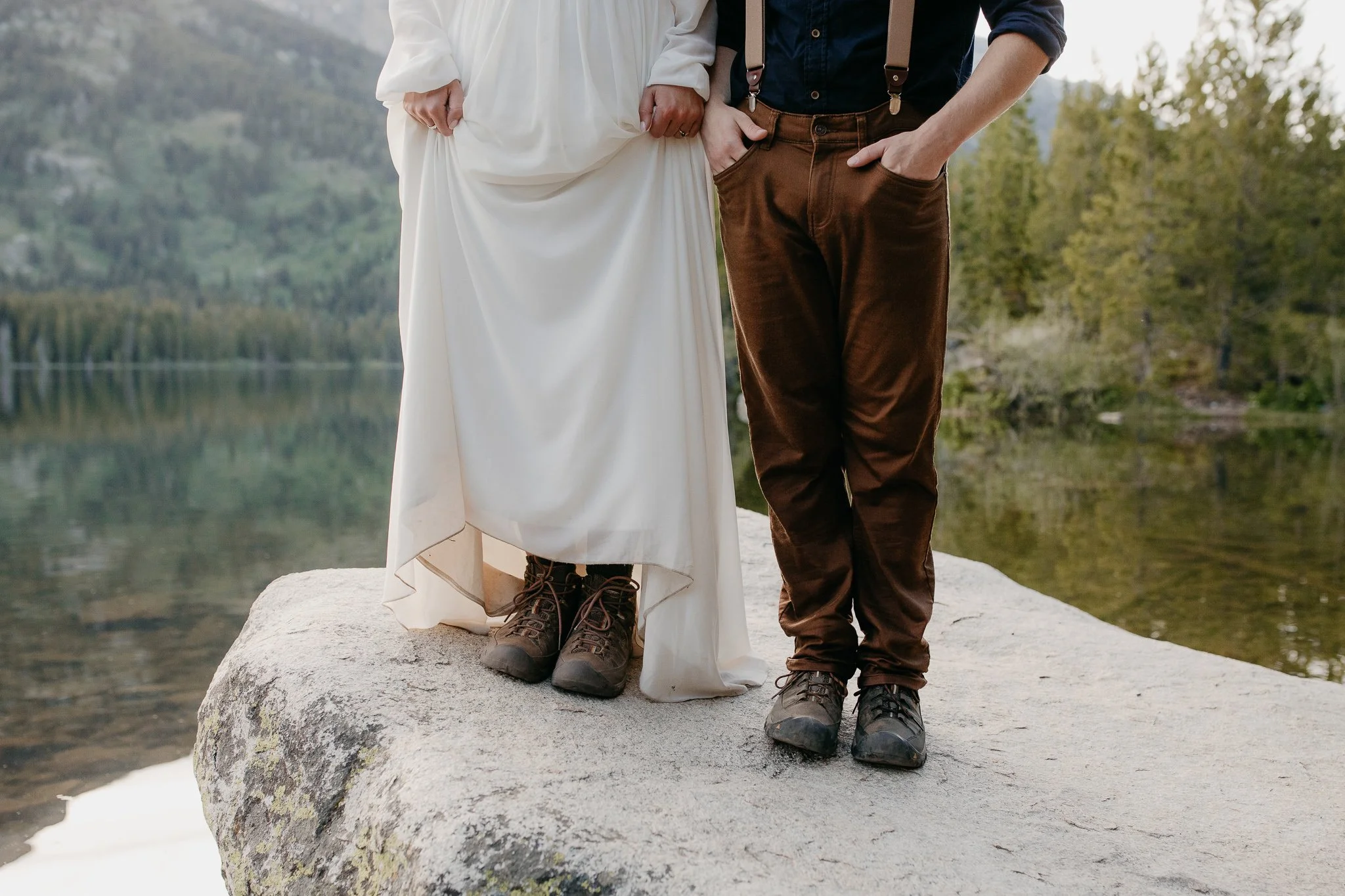 Bride and groom standing on a rock by a lake wearing hiking boots during their elopement