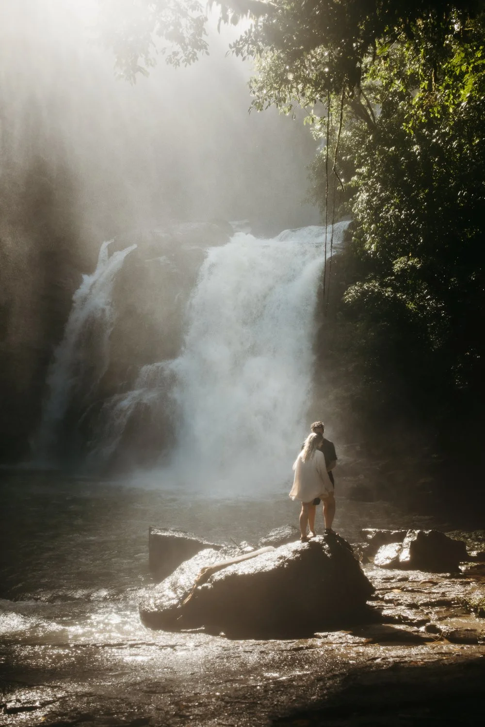 Couple standing on rocks in front of a waterfall in a lush natural landscape