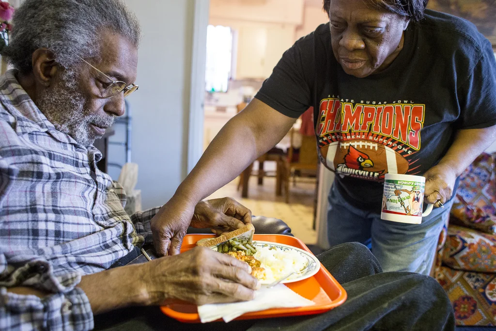 Beatrice Ford serves her husband, Hugh, a meal of part Detroit Meals on Wheels delivered food and part her own cooking in their home, April 18, 2014. She serves it to him on a red plastic tray and balances a lamp on a TV table to help him see.