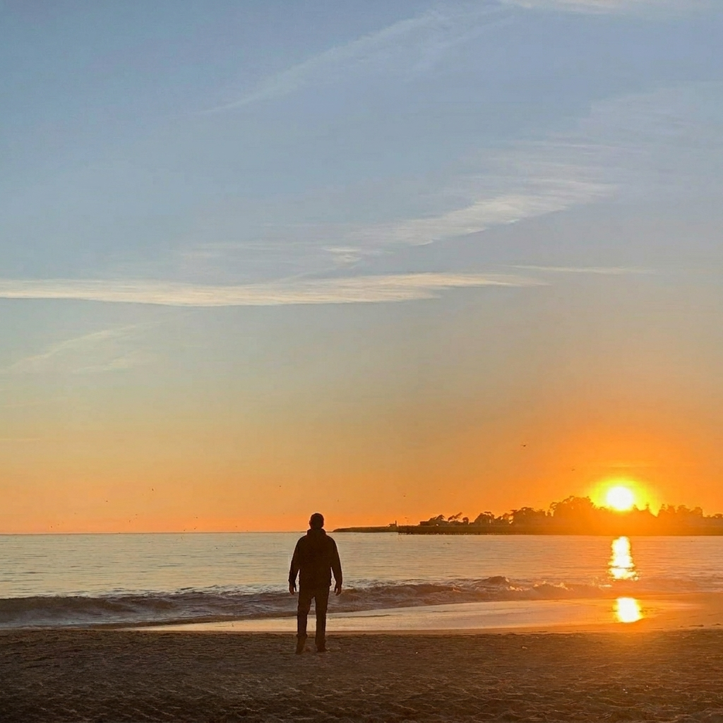 A person standing on the beach during sunset, with the sun low on the horizon over the water, and a sky with clouds overhead.