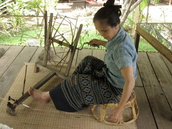 Cotton spinning in Laos, using a hand-turned driven spindle wheel (and recruiting the foot to hold the wheel in place.) (Ock Pop Tok Living Crafts Centre, Luang Prabang, 2013)