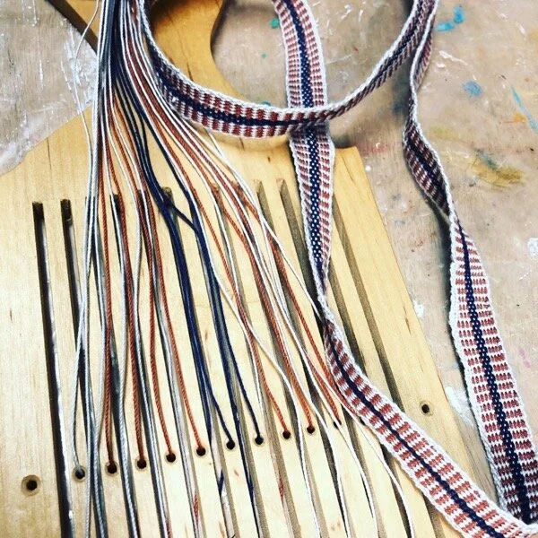Wooden “knee heddle” with crochet cotton tape being woven in stripes of rust and grey, with blue down the middle. This image shows how the warps are closer together in one section, which I’d prefer for the whole length.