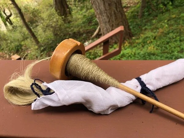 Spindle with flax cop and flax fiber folded and tied into a hankie. Green plants and trees in background.
