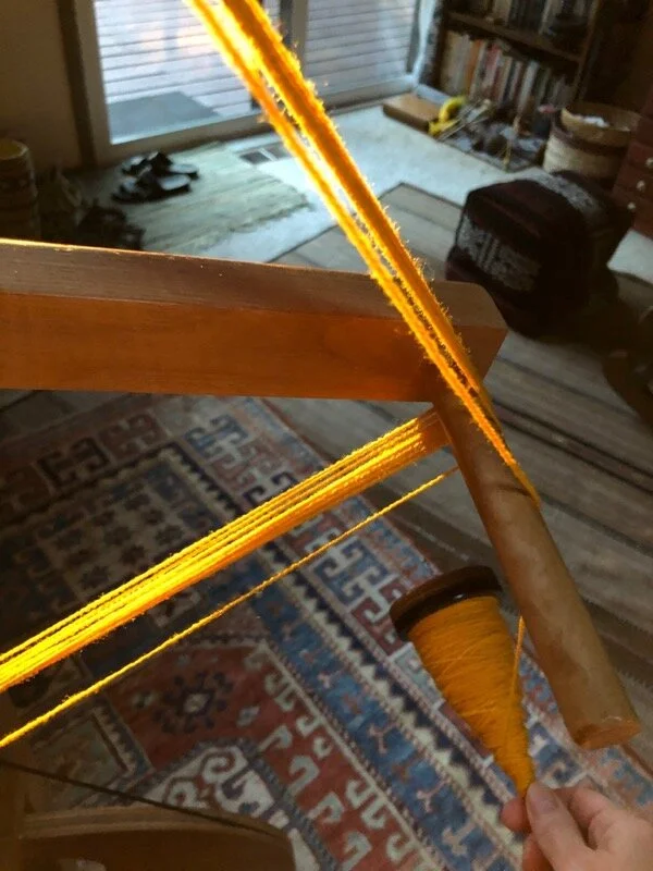 Winding sunlit yellow handspun yarn off a spindle onto a reel, Afghani and Bedouin rugs in the background.