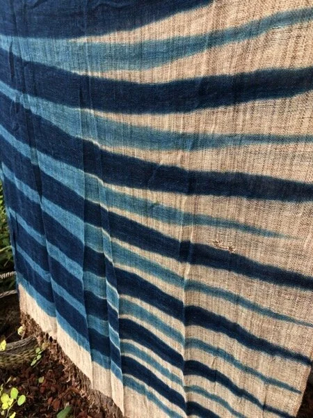 Large scarf made of plant fiber dip-dyed with tapered horizontal stripes of indigo in alternating light and dark tones. Creased from being folded….