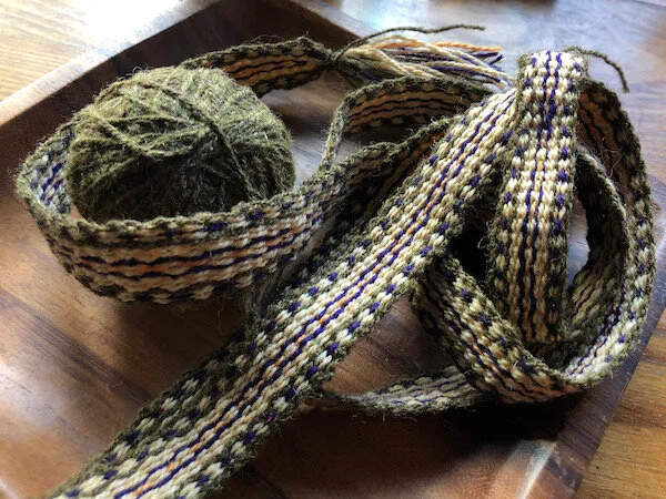 Spindles-spun wool in heathered green, deep purple, pale orange and bright orange with warped-in design. Ball of green handspun wool.