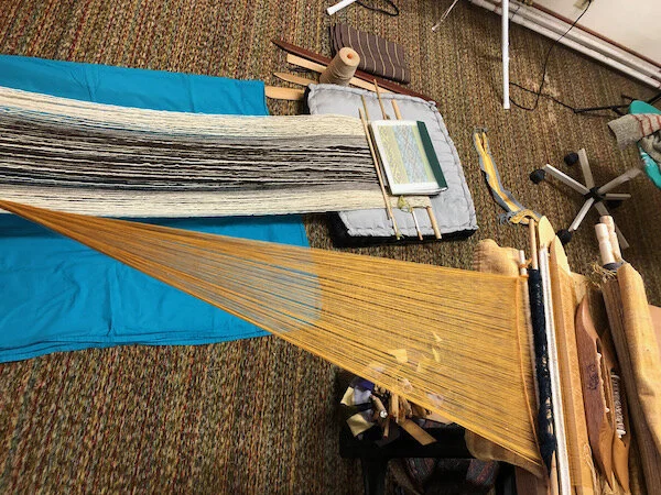 Double decker weaving. When you have one tie-up spot for larger pieces, they have to make way for one another. The gold warp is chained and secured at one point, so it can move side to side. I weave it while sitting in the rolling chair. The wool warp is secured on a loom bar, so it faces the tie-up (antique treadle sewing machine, that is) directly, and I sit on a cushion on the floor to weave it.