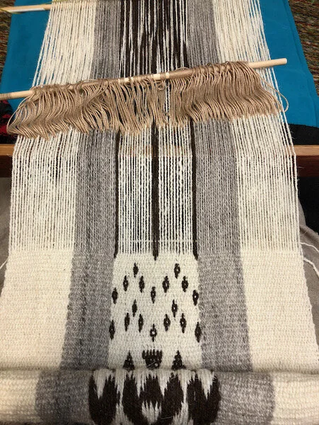 Woven cloth with pickup design in the middle, grey stripes to either side, white borders that will join the white of the side panels. String heddles and shed stick behind. Heddled shed is open, design is picked up.