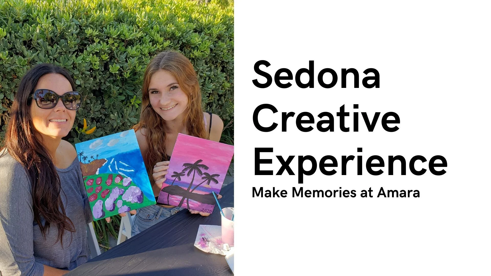 Two women at an outdoor art event holding colorful landscape paintings, with greenery in the background and a table with painting supplies.
