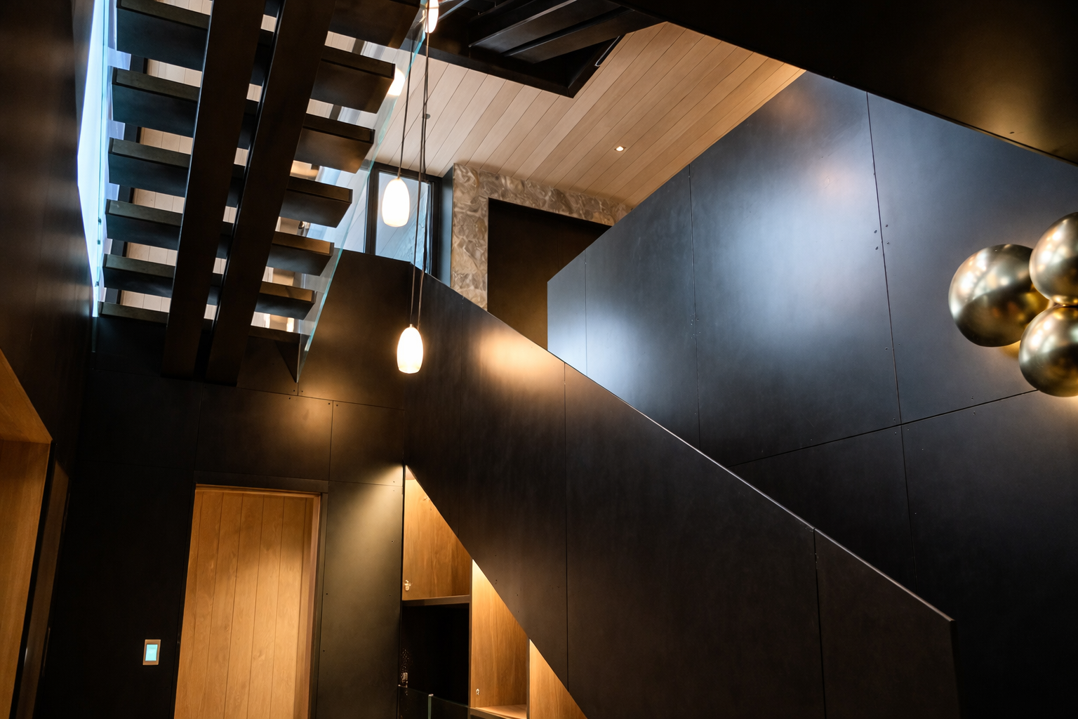 Modern interior staircase with black metal and wood elements, pendant lights, and black walls surrounding a wooden door.