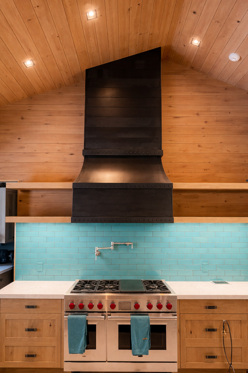 Modern kitchen with wooden ceiling, black range hood, teal subway tile backsplash, and stainless steel stove with red knobs.