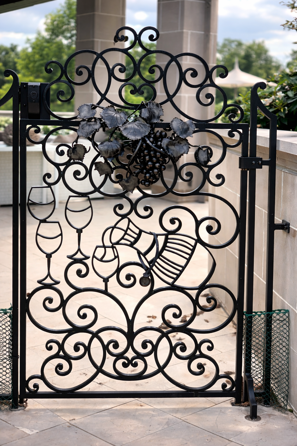 Decorative black metal gate with grapevine and wine glass motifs, situated outdoors on a patio with trees in the background.