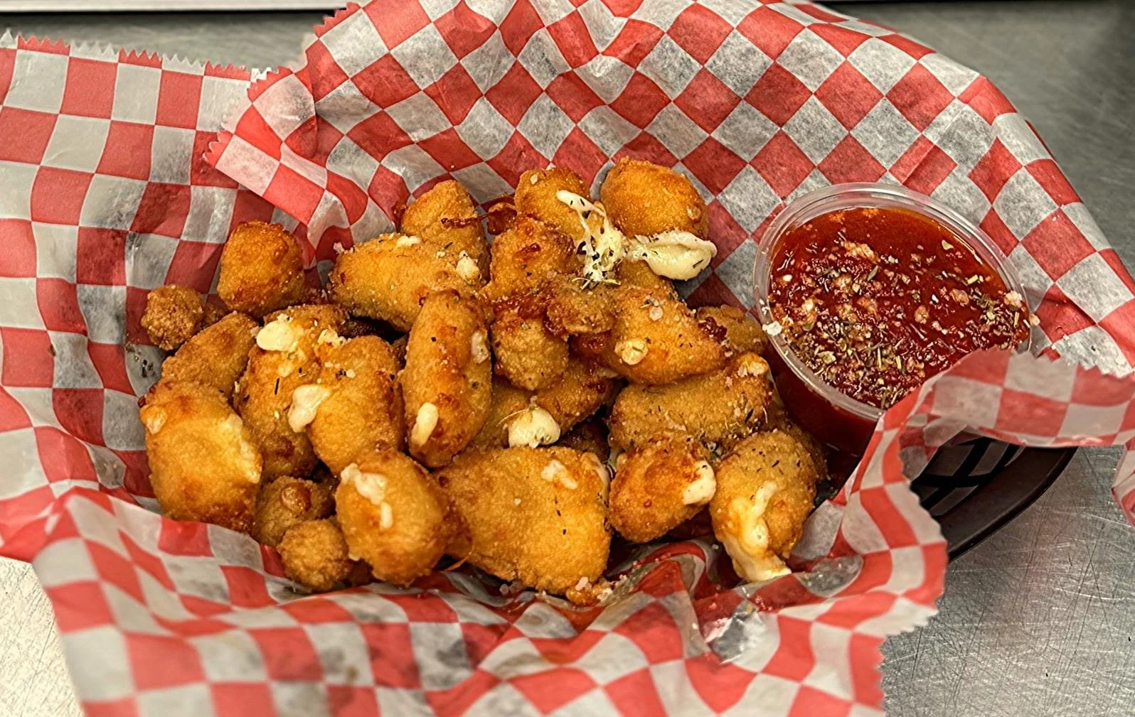 Fried cauliflower bites with garlic and cheese, served with a cup of red dipping sauce on red and white checkered paper in a basket.