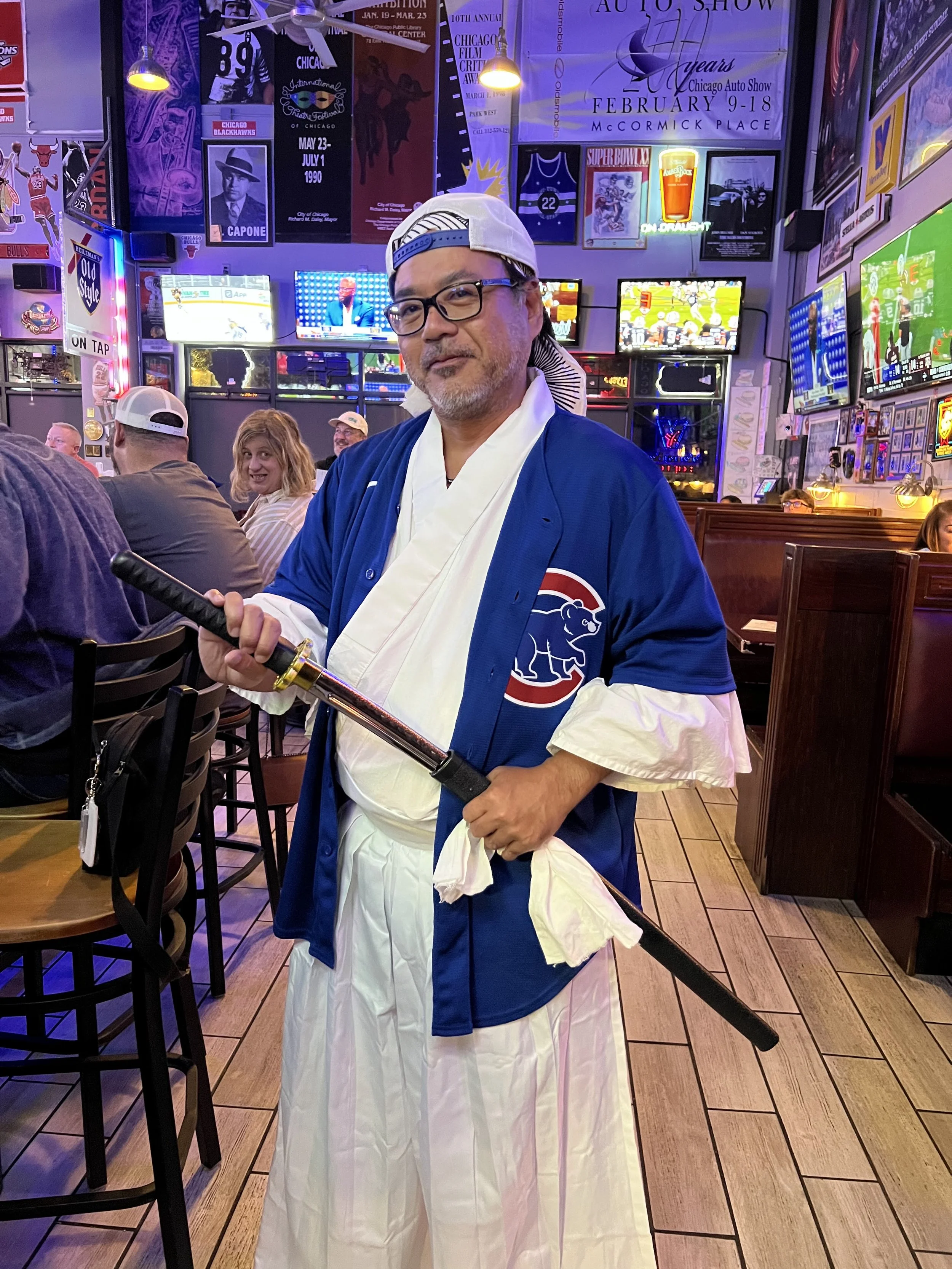 A man dressed in a Chicago Cubs baseball jersey, traditional Japanese clothing, and a baseball cap, holding a katana sword inside a sports bar with multiple television screens showing sports games.