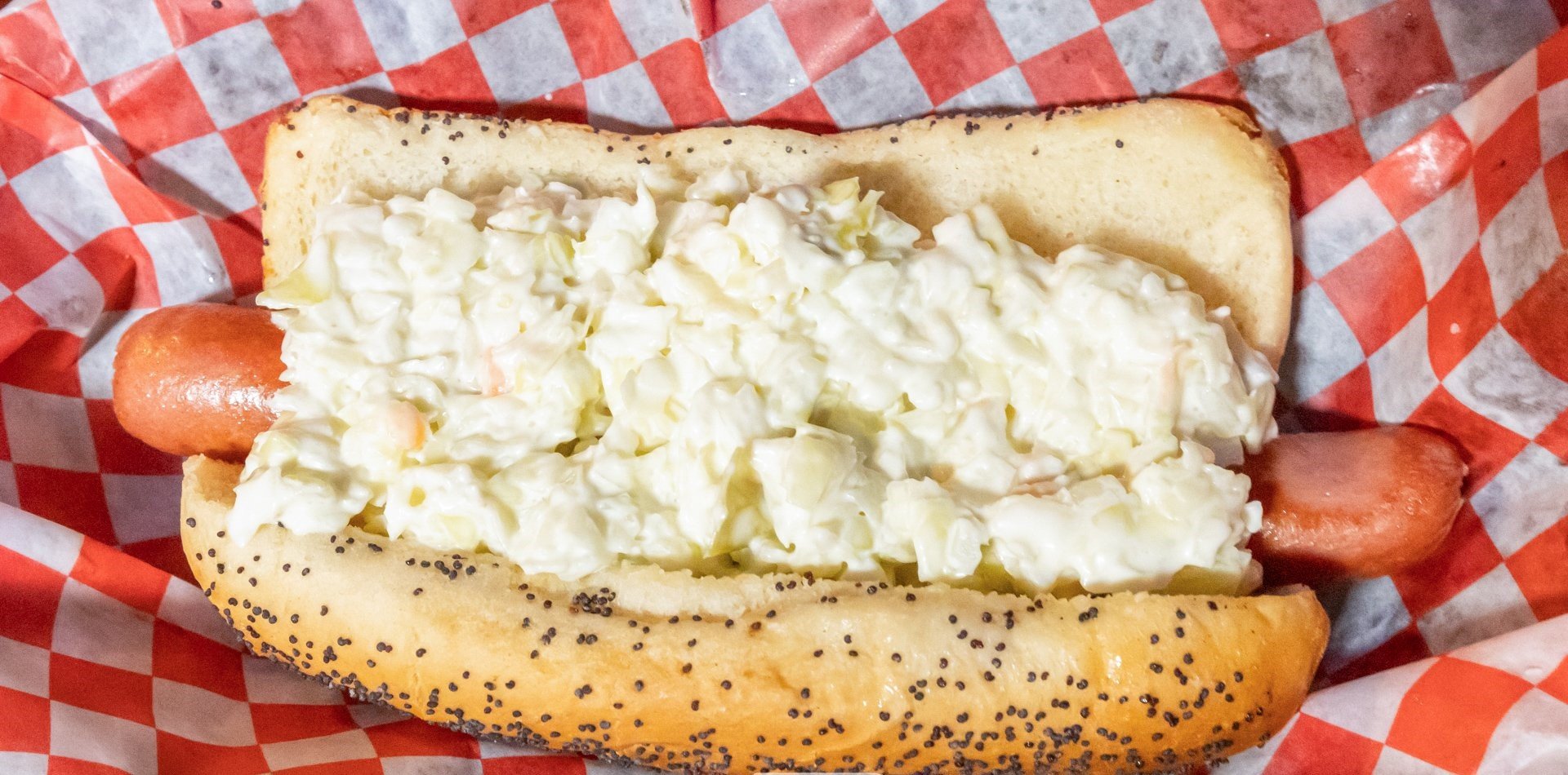 Hot dog in a poppy seed bun with chopped egg salad inside, resting on red and white checkered paper.