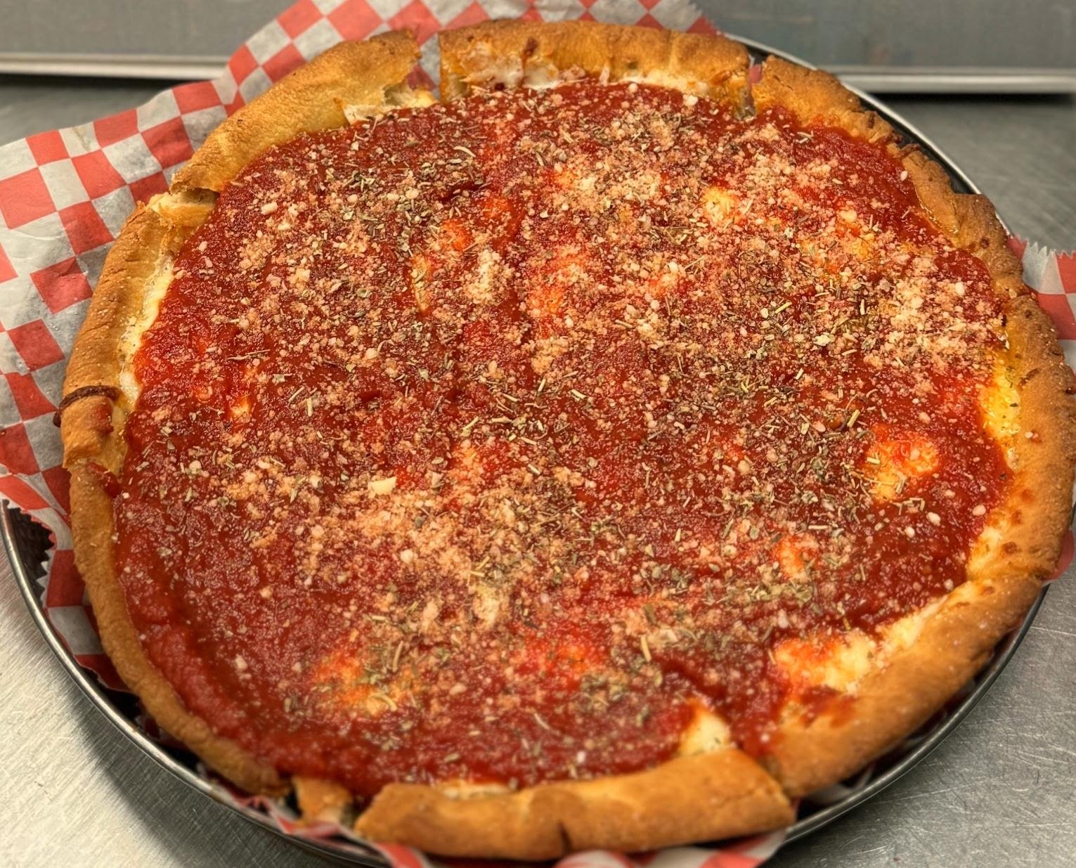 A large, round pizza with a thick crust, topped with tomato sauce and sprinkled with herbs and grated cheese, sitting on red and white checkered paper in a metal pan.