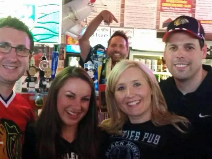 A group of five friends smiling and posing for a photo in a bar or sports pub, with a man in the background making a gesture with his hand.