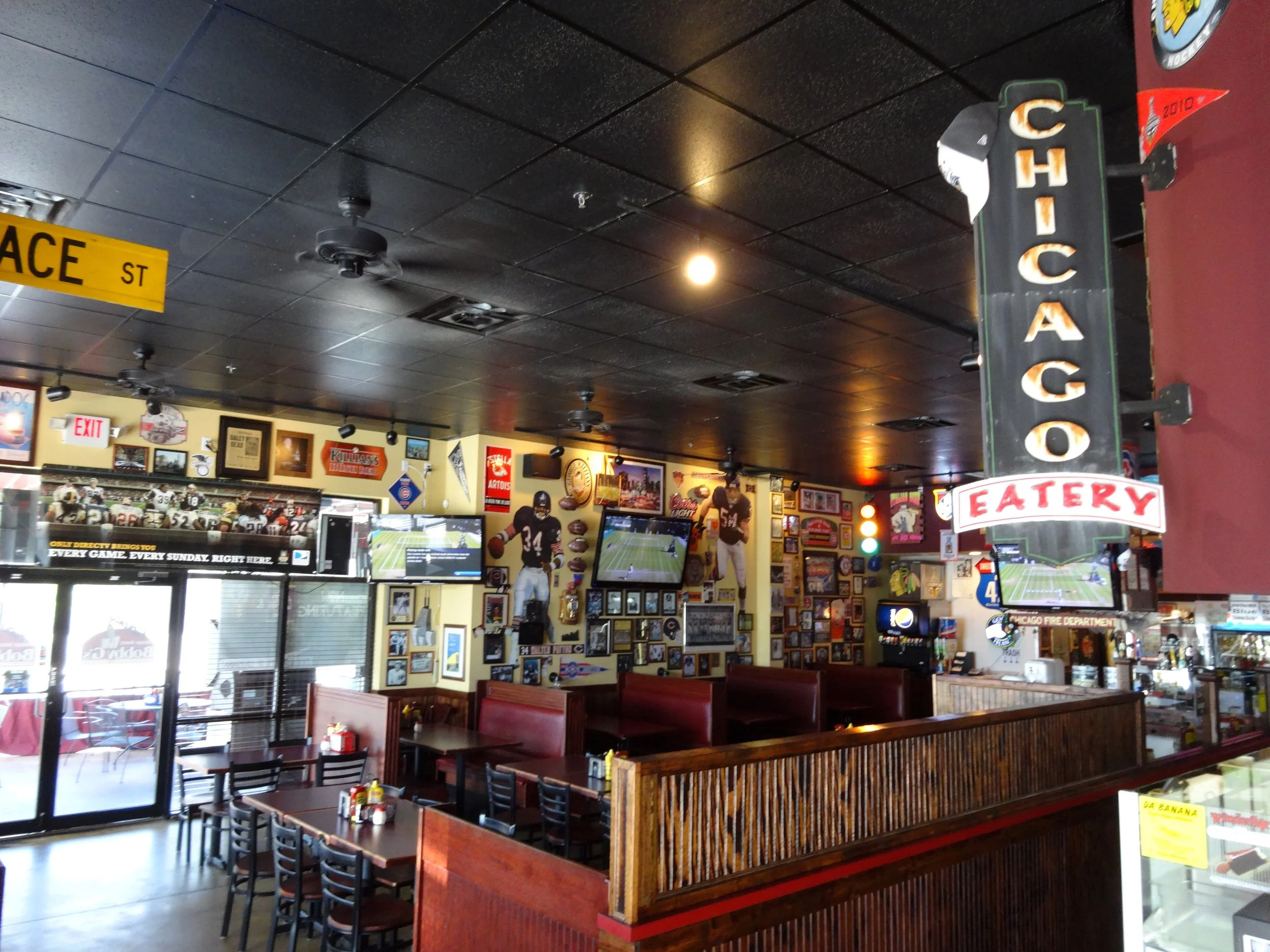 Interior of a sports bar with multiple televisions showing football and walls decorated with sports memorabilia, jerseys, and framed pictures. There are tables with chairs and condiments, and a neon sign reading 'Chicago Eatery' hanging from the ceil