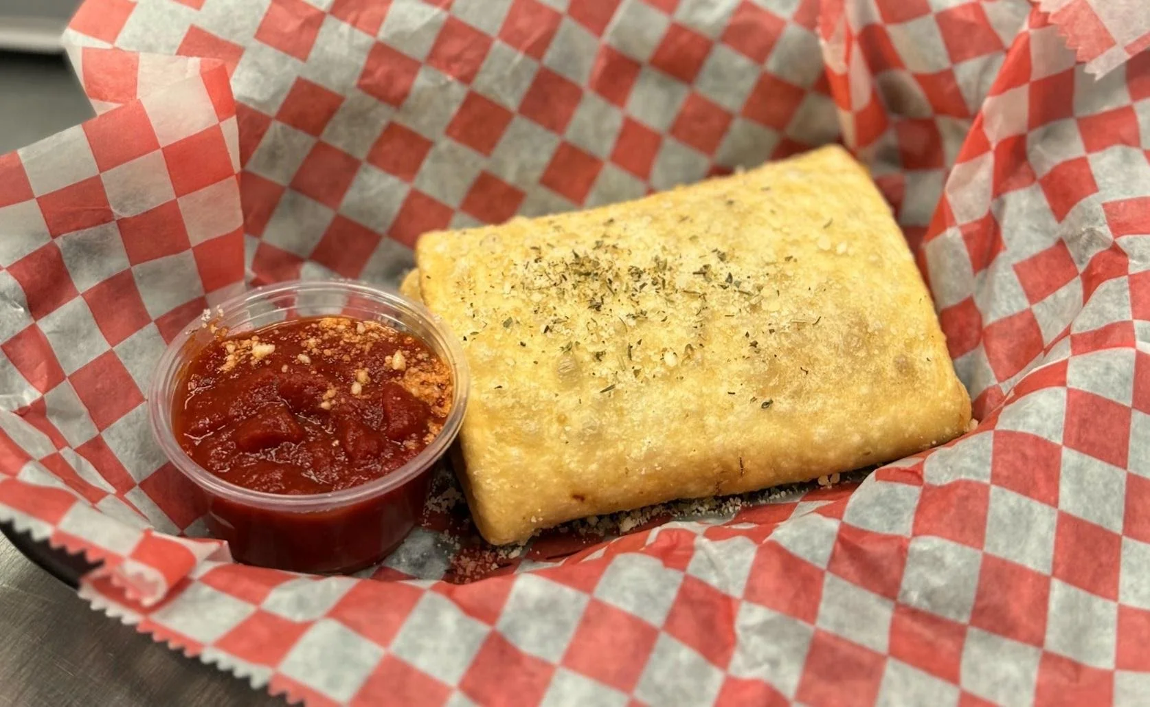 A piece of fried bread topped with herbs, served with a cup of marinara sauce on red and white checkered paper in a basket.