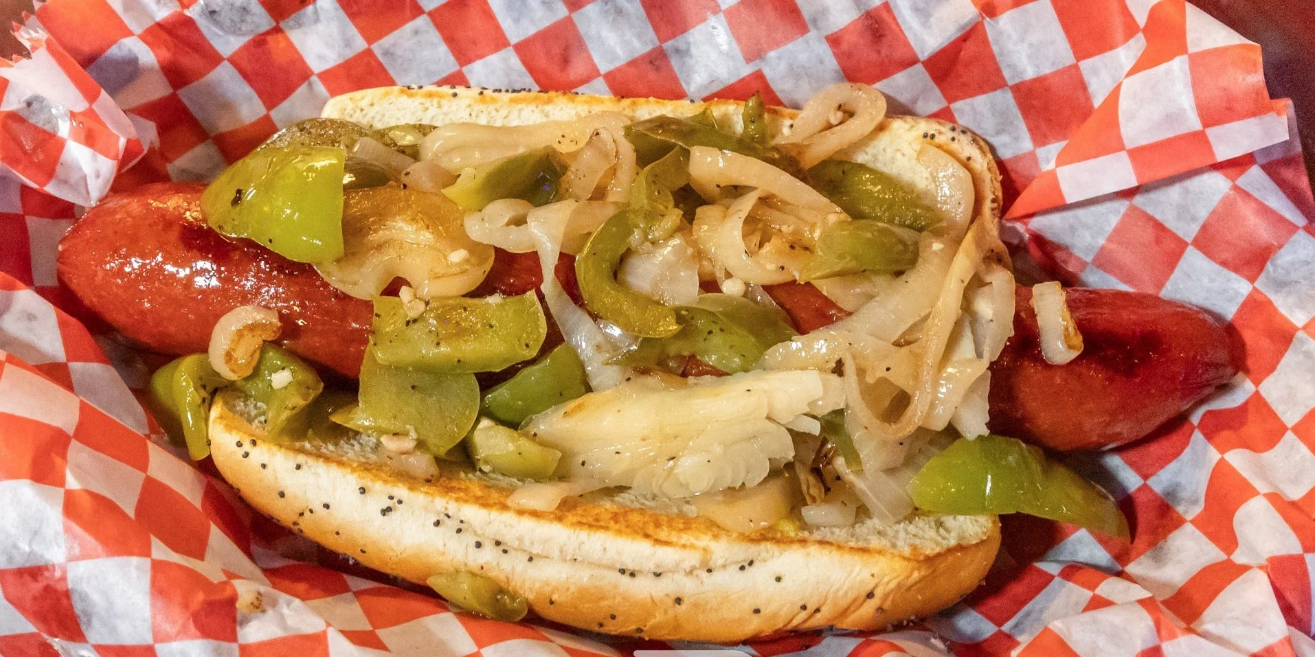 Hot dog with grilled sausage in a poppy seed bun topped with sautéed onions and green bell peppers.