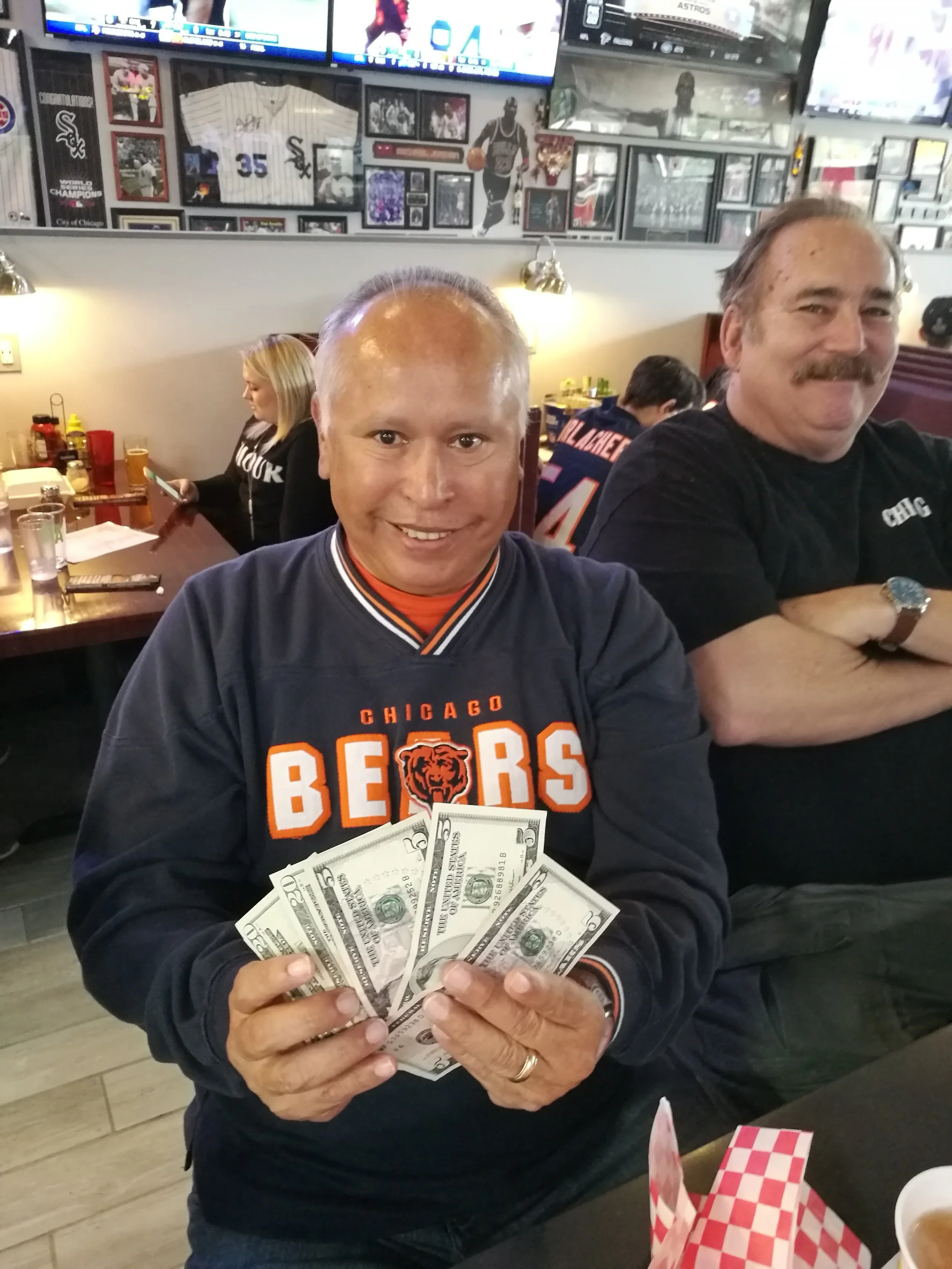 A man wearing a Chicago Bears sweatshirt holding several five-dollar bills in a restaurant or bar.