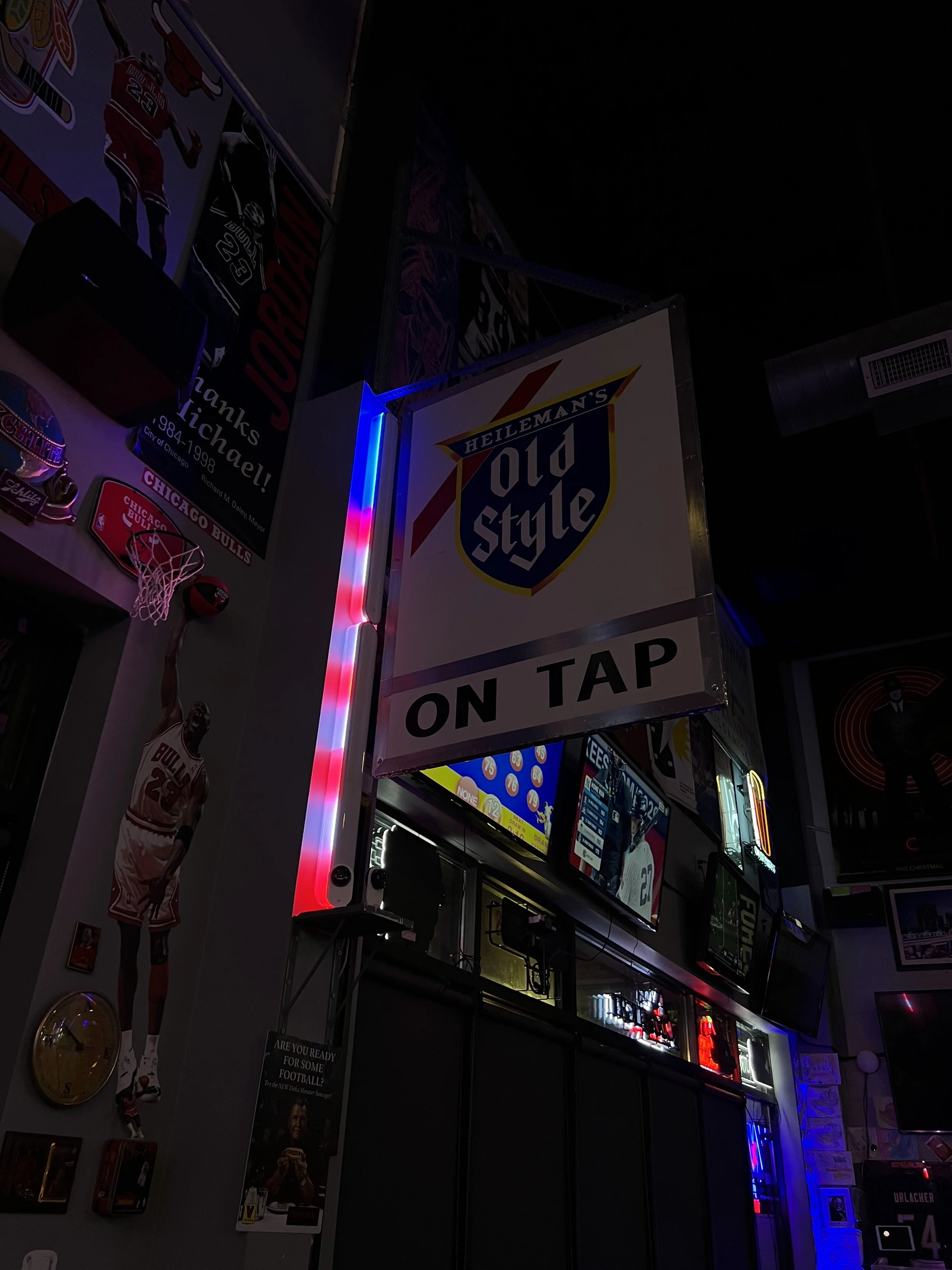 Indoor scene of a bar or sports game venue with multiple television screens, sports posters, and a sign for Old Style beer that reads 'ON TAP'.