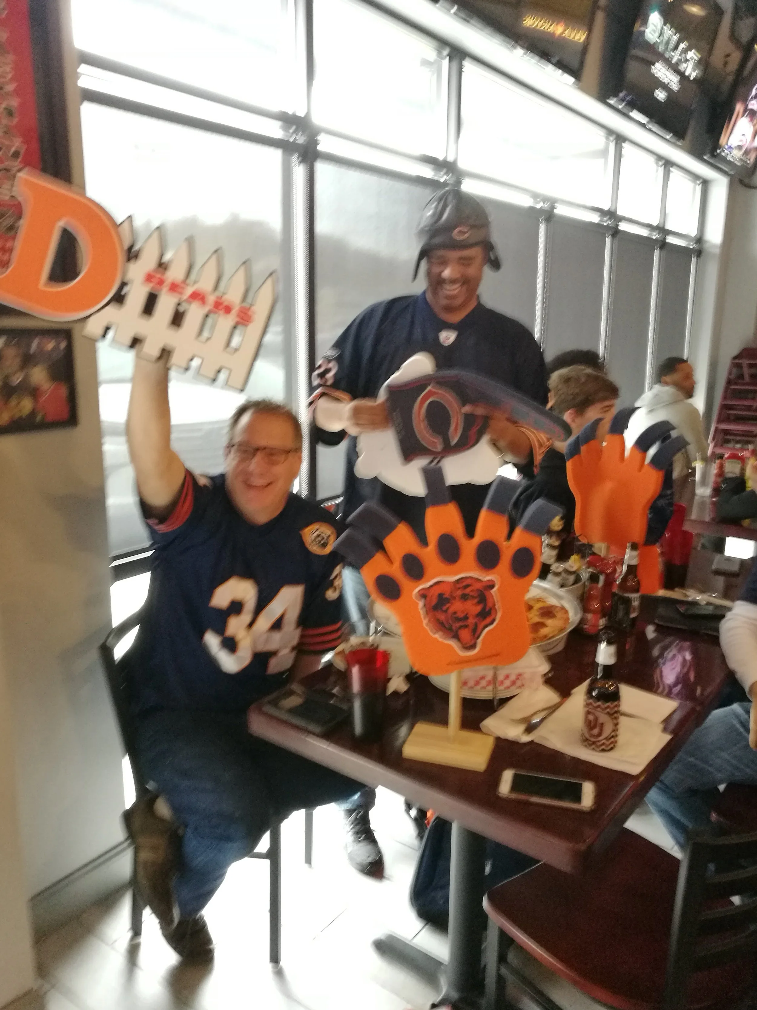 Two men celebrating at a Chicago Bears-themed gathering, wearing football jerseys, with decorations including foam fingers and Bears logos on a table, in a restaurant or bar setting.