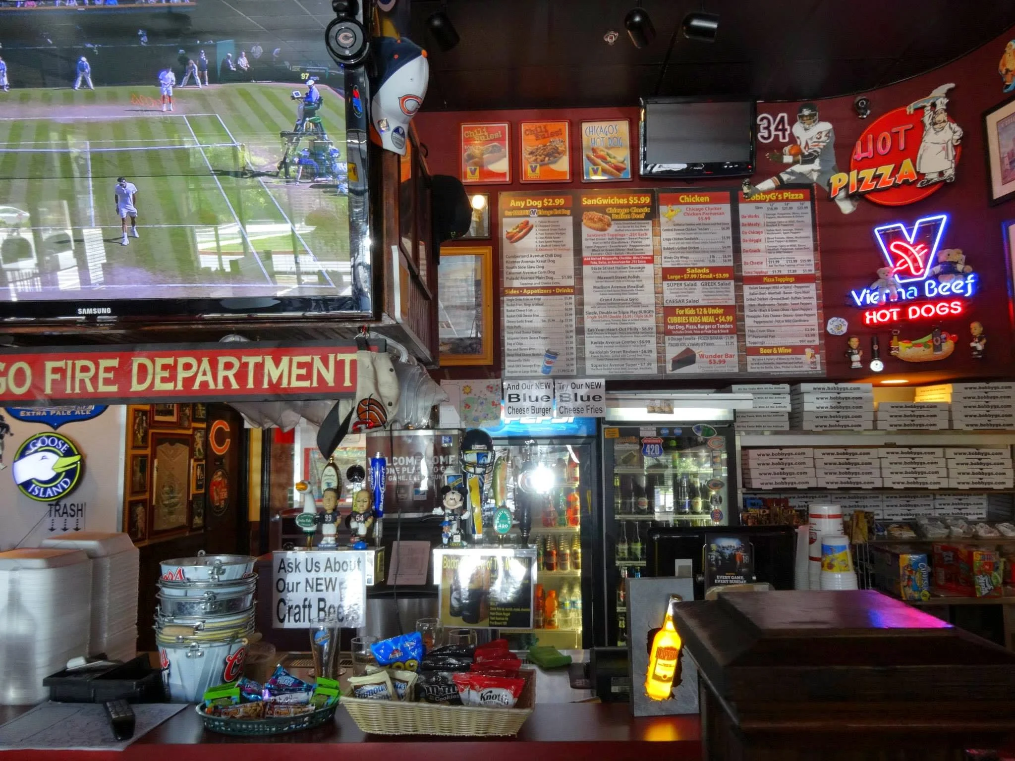 Interior of a sports bar featuring a large TV showing a tennis match, colorful menu boards, neon signs, and a counter with snacks and condiments, including a sign inviting customers to ask about craft beers.