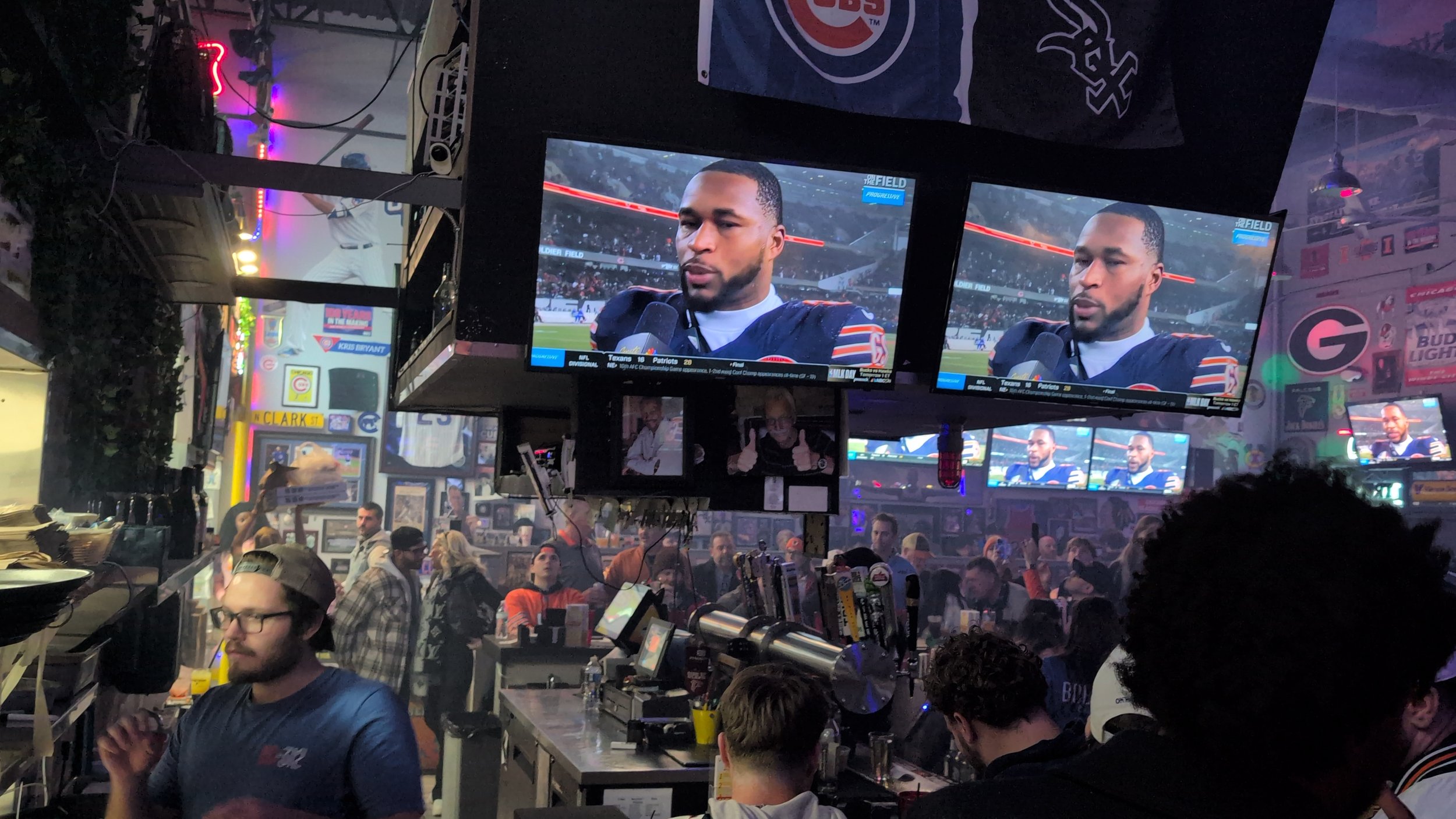 A sports bar showing a football game on multiple large TVs. One TV features a close-up of a football player giving an interview, with fans watching and socializing in the background.