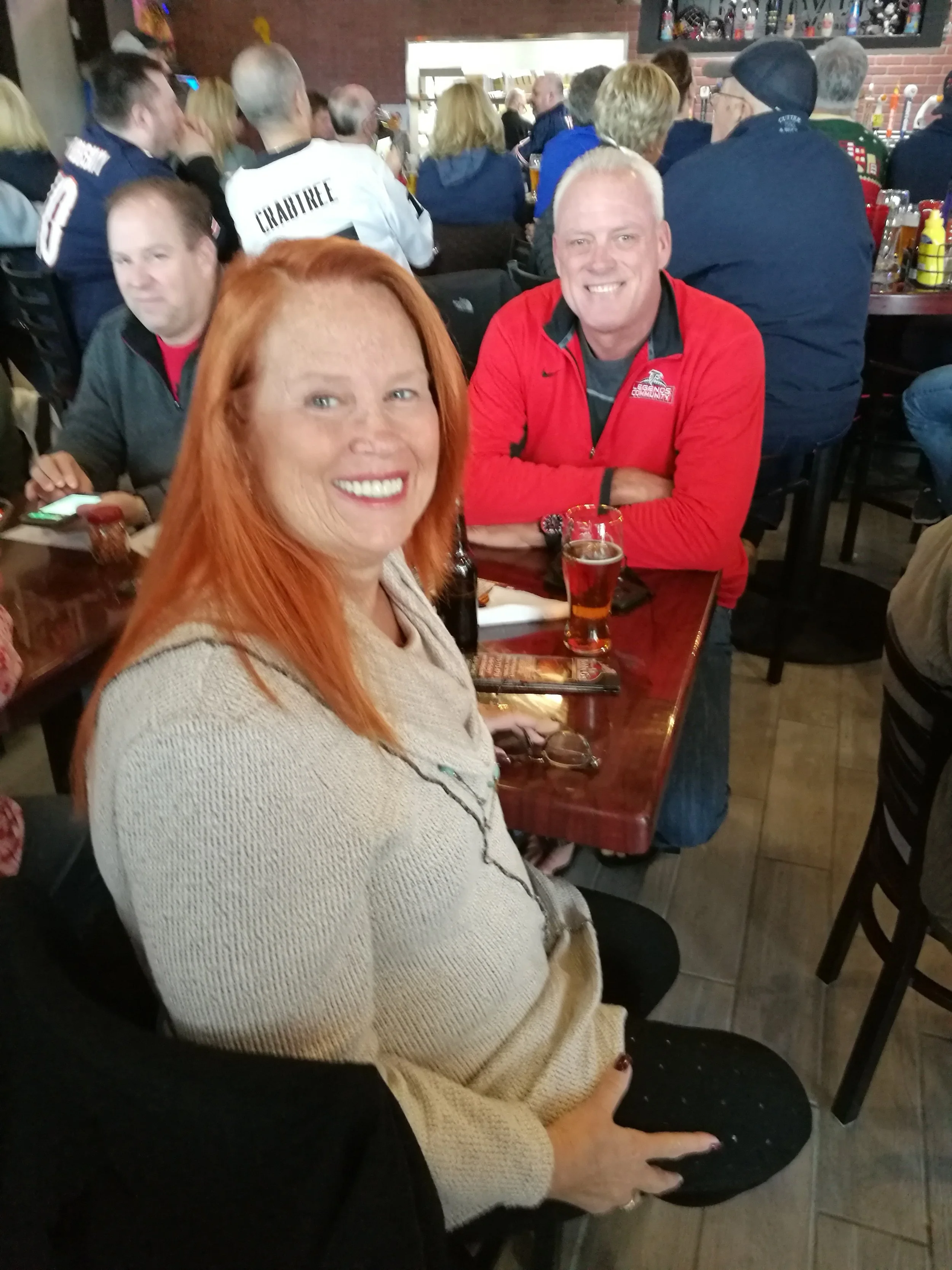 A woman with red hair smiling, sitting at a table in a lively bar or restaurant, with a man in a red jacket smiling in the background, surrounded by other patrons.